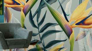 A gray armchair sits by a wall adorned with vibrant, abstract tropical leaves and flowers in hues of orange, yellow, green, and purple. The scene is modern and lively.