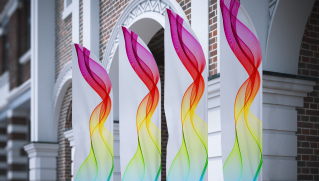 Four white vertical banners with vibrant rainbow wave patterns stand in front of a brick building with arches, creating a lively and modern look.